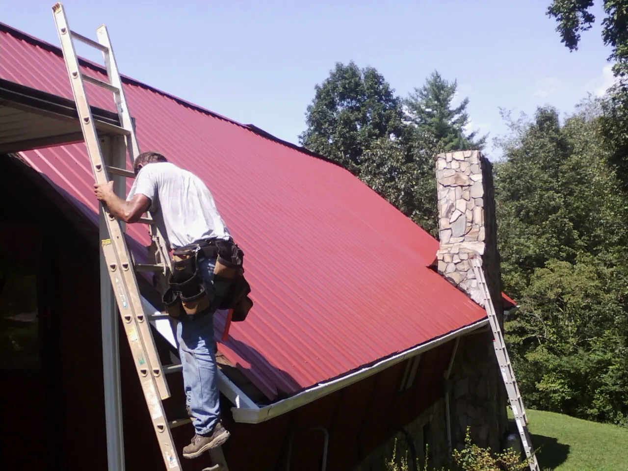 Roofer on a ladder beside a red metal roof and stone chimney — Soffit & Fascia Repair by Roofing Brothers in Elgin